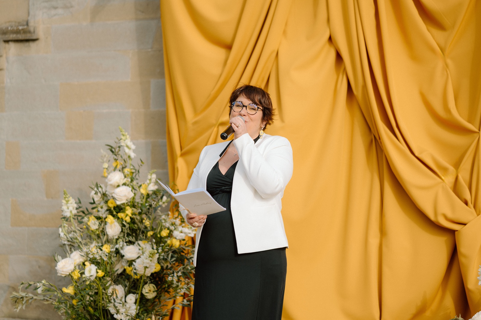 Alice — chant pour une cérémonie de mariage Alice chante au micro, un livret à la main, devant un drapé doré et une composition florale.