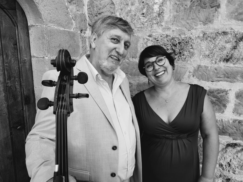Portrait en noir et blanc d’Alice et d’un violoncelliste échangeant un sourire devant un mur de pierre.